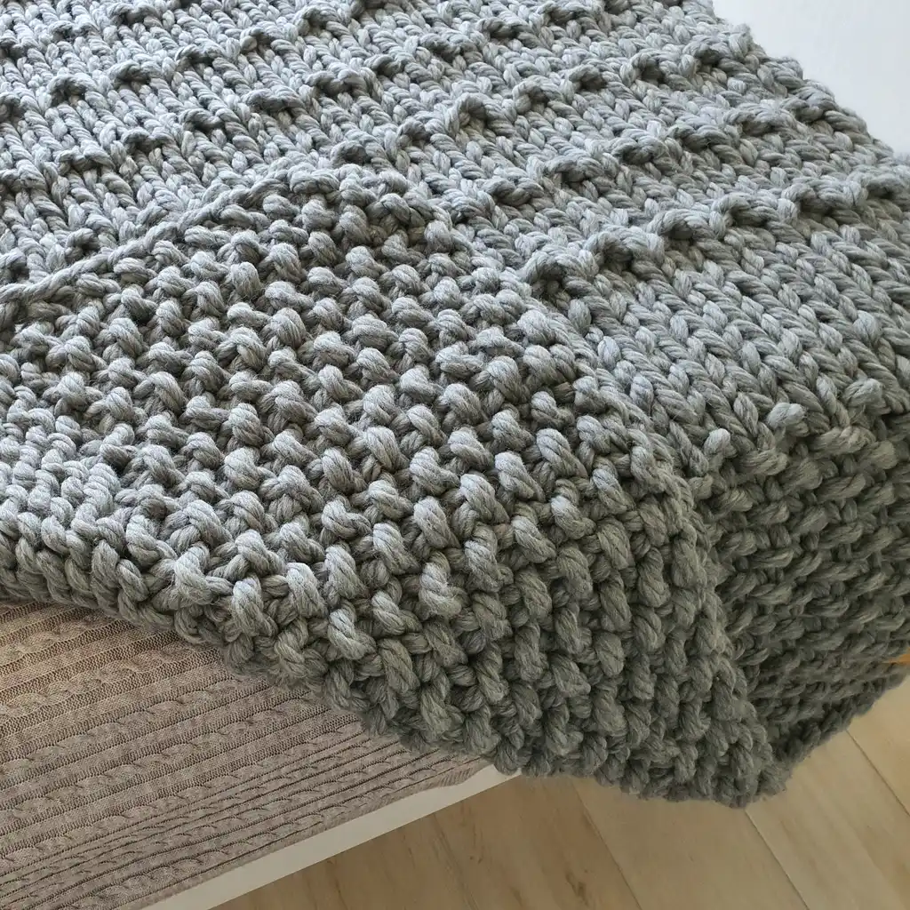 Chunky wool knitted blanket in neutral color with textured pattern, displayed on modern furniture.