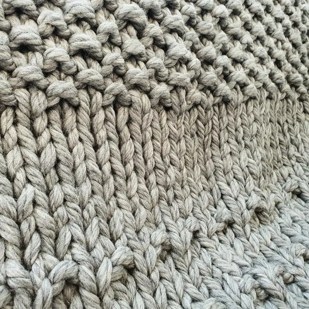 Chunky wool knitted blanket in neutral color with textured pattern, displayed on modern furniture.