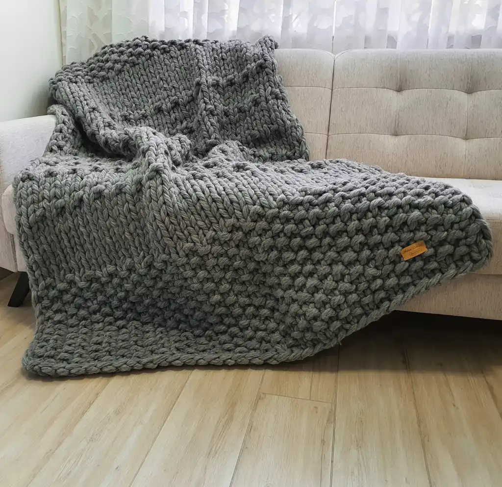 Chunky wool knitted blanket in neutral color with textured pattern, displayed on modern furniture.