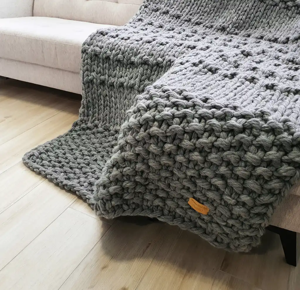 Chunky wool knitted blanket in neutral color with textured pattern, displayed on modern furniture.