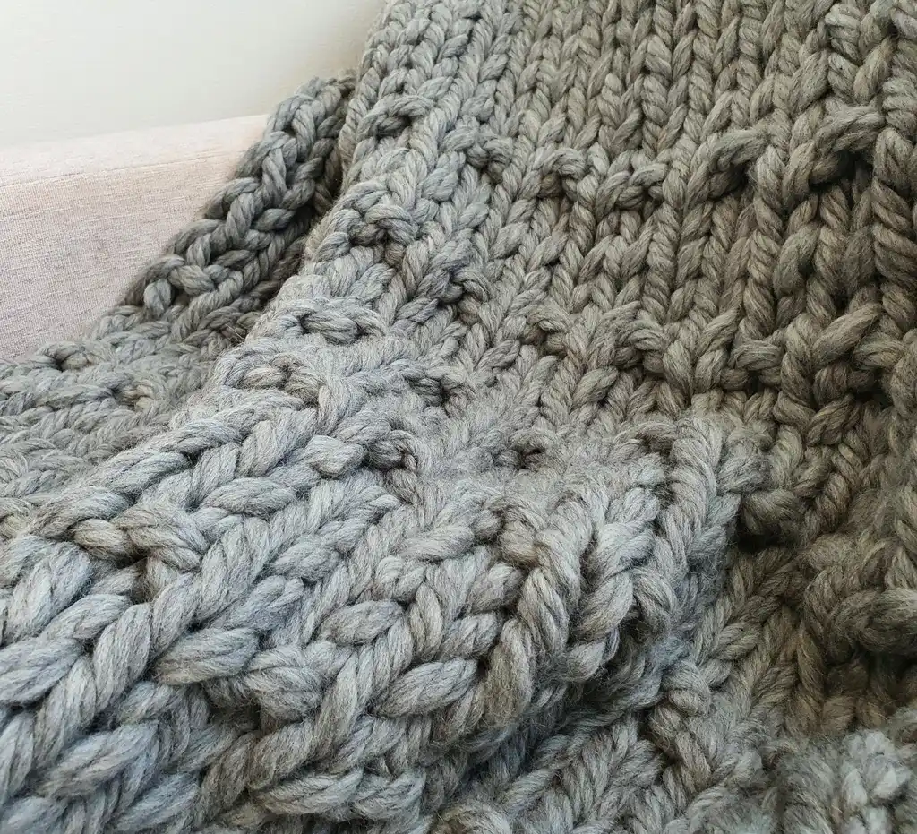 Chunky wool knitted blanket in neutral color with textured pattern, displayed on modern furniture.