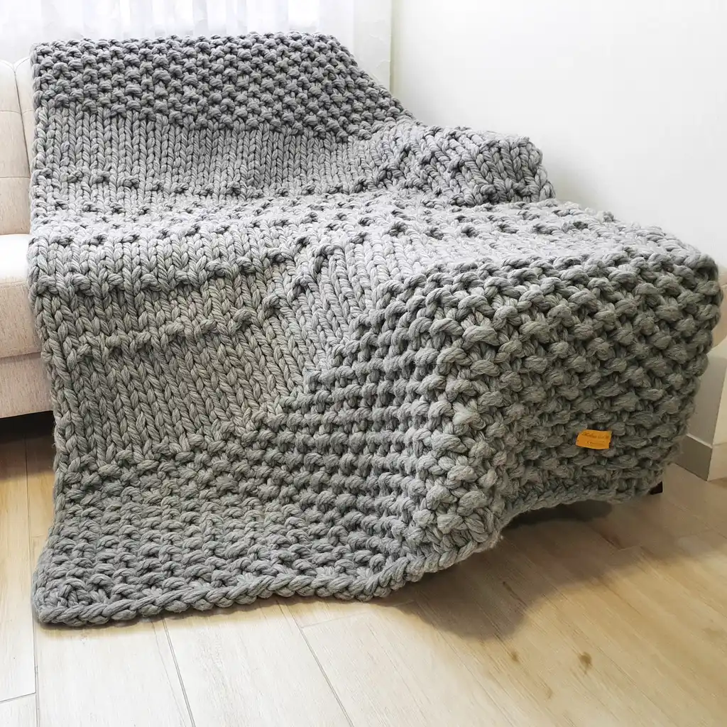 Chunky wool knitted blanket in neutral color with textured pattern, displayed on modern furniture.