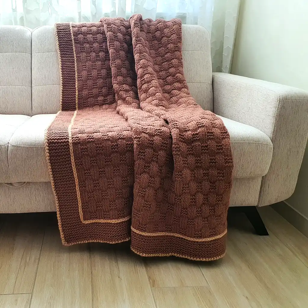 Handmade chunky knitted throw in terracotta color with beige border, textured pattern.