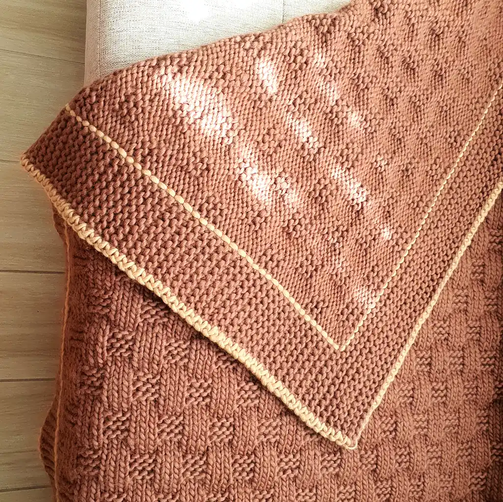 Handmade chunky knitted throw in terracotta color with beige border, textured pattern.
