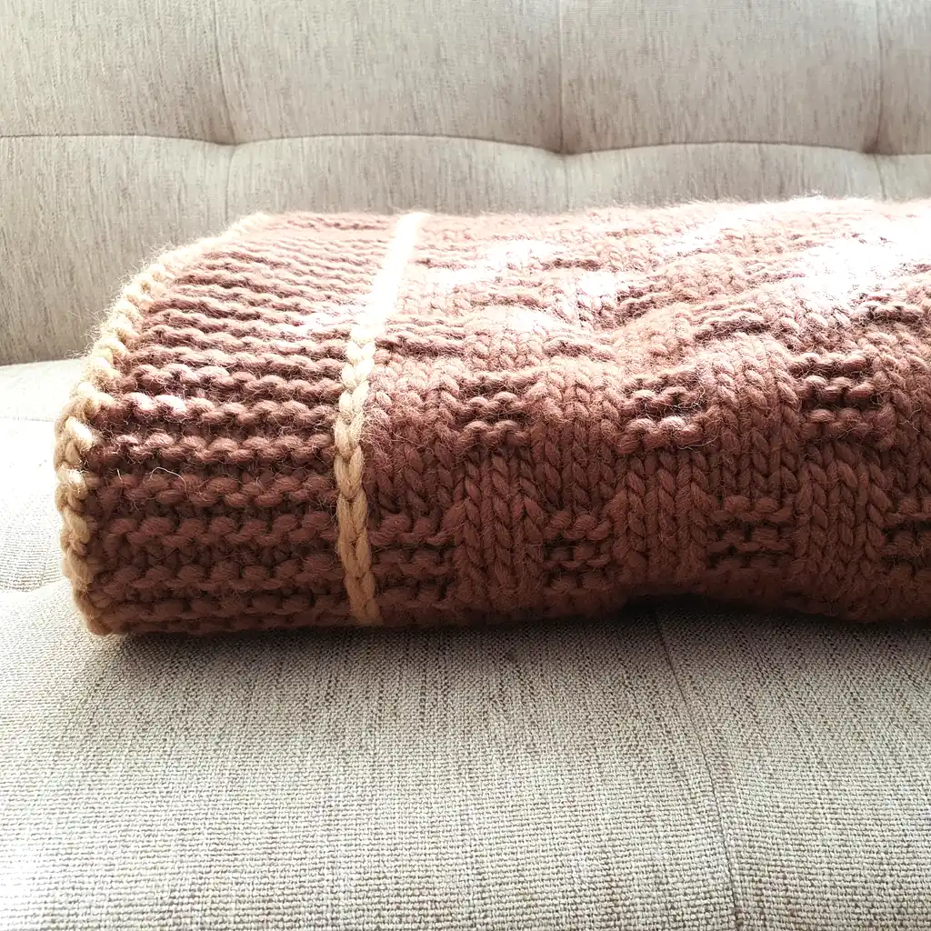 Handmade chunky knitted throw in terracotta color with beige border, textured pattern.