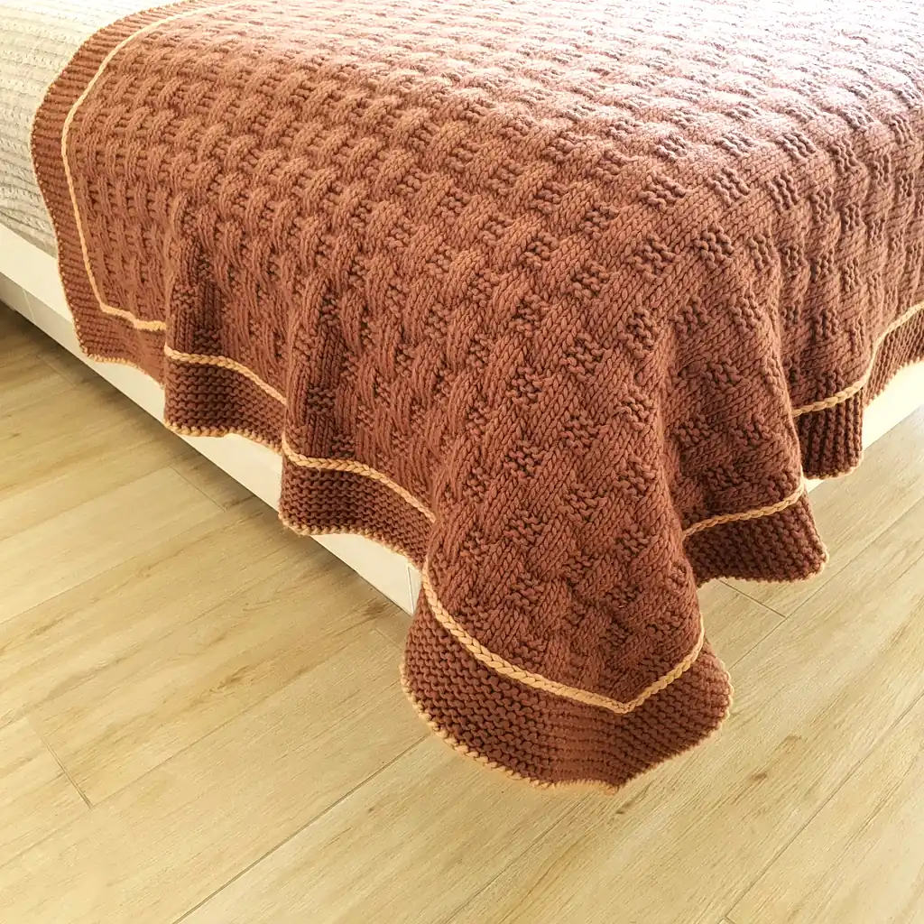 Handmade chunky knitted throw in terracotta color with beige border, textured pattern.