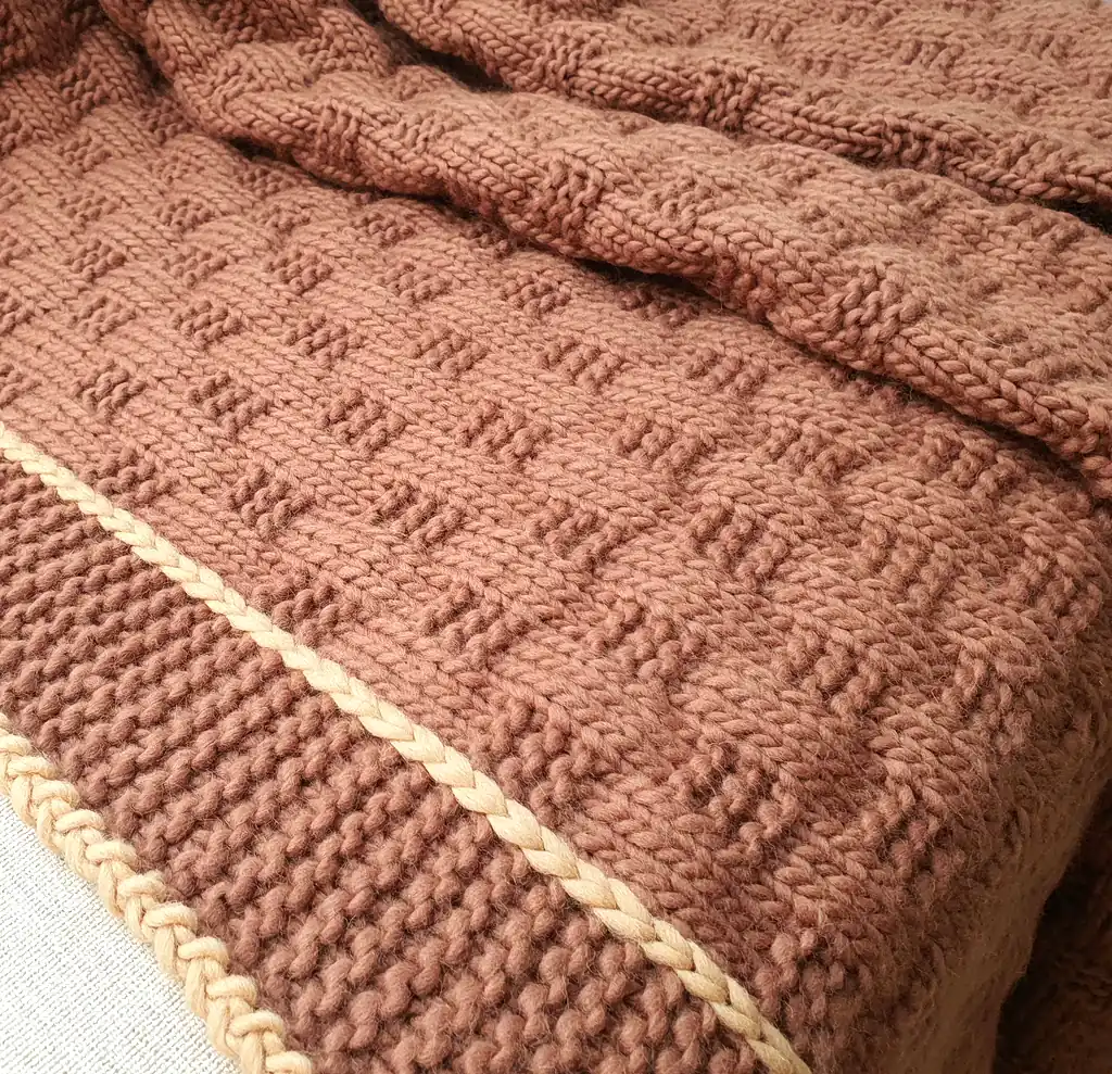 Handmade chunky knitted throw in terracotta color with beige border, textured pattern.