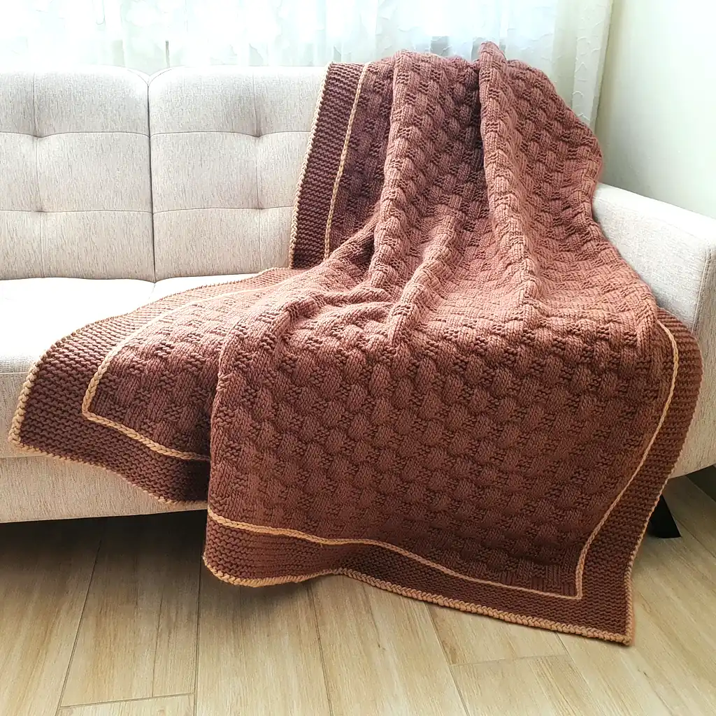 Handmade chunky knitted throw in terracotta color with beige border, textured pattern.