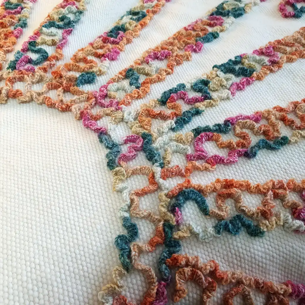 Close-up of textured embroidery in orange, green, and pink on soft cream wool fabric.
