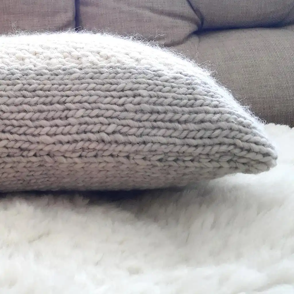Handmade merino wool knitted cushion with textured pattern in gray and warm white colors.